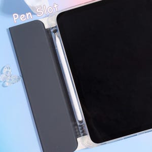 Pode incluir: Uma capa transparente para tablet com tela preta e caneta branca. A capa tem uma capa cinza escuro com as palavras "Pen Slot" impressas na parte superior. Uma decora&ccedil;&atilde;o de borboleta transparente &eacute; vis&iacute;vel no lado esquerdo.