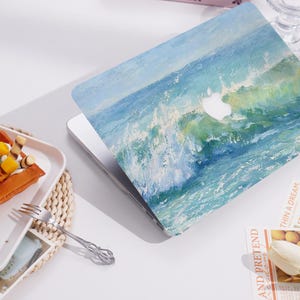 May include: A laptop with a cover depicting an ocean wave painting in blue and green hues. The Apple logo is visible. The laptop sits on a white surface, with a waffle and fork in the foreground.