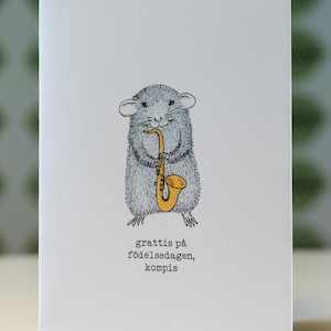 May include: A white greeting card with a cartoon illustration of a grey mouse playing a yellow saxophone. The text "grattis på födelsedagen, kompis" is printed below the mouse. The card has a simple, whimsical design.