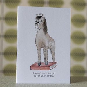 May include: Greeting card featuring a whimsical illustration of a horse wearing glasses and a hat, standing on a book. The card has text at the bottom in a foreign language. The background is a pattern of green and yellow circles.