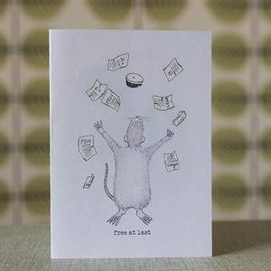 May include: A white greeting card with a black and white illustration of a rat with its arms raised, surrounded by falling papers and a hat. The words "free at last" are printed at the bottom.