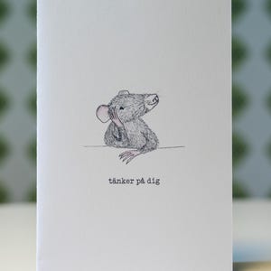 May include: A white card with a hand-drawn illustration of a grey mouse, its paw on its head. The text "tänker på dig" is printed in black below the mouse.
