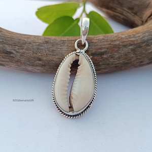 May include: A silver pendant featuring a white cowrie shell with a detailed silver border. The pendant is on a wooden surface.