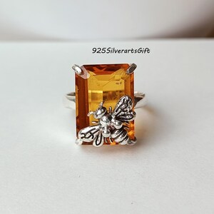 Puede incluir: Anillo de plata con una gran gema rectangular de color ámbar. Una abeja plateada detallada está unida a la gema. El texto "925SilverartsGift" es visible.