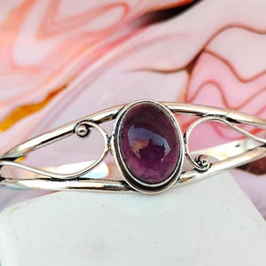 Può includere: Un braccialetto d'argento con una pietra preziosa viola incastonata al centro. Il braccialetto ha un design delicato e ornato.