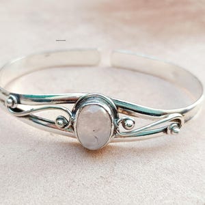 Könnte beinhalten: Ein silbernes Armkettchen mit einem filigranen Design, das einen weißen ovalen Edelstein zeigt. Das Armband hat einen schlichten, eleganten Stil.