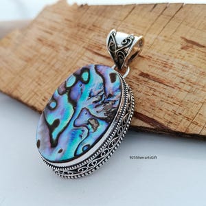 May include: A silver pendant with an oval abalone shell inlay. The shell has iridescent blue, green, and purple colors. The pendant has a detailed silver border and a bail for attaching a chain.