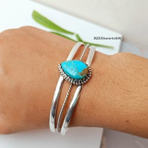 May include: Silver cuff bracelet with a turquoise stone. The stone is teardrop-shaped, set in a beaded bezel, and framed by two parallel silver bands and a twisted silver rope. The text "925silverartsGift" is visible.