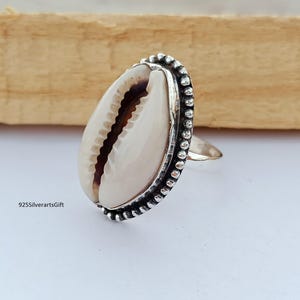 Op de afbeelding: Zilveren ring met een grote, crèmekleurige kaurischelp. De schelp is gezet in een decoratieve zilveren band met een kralenrand. De ring heeft een eenvoudig, elegant ontwerp, perfect voor een vleugje kuststijl.