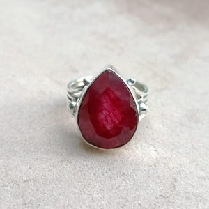 May include: A silver ring featuring a large, teardrop-shaped ruby gemstone. The ruby is a deep red colour and has a faceted cut.