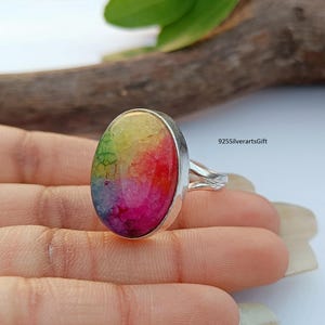 Puede incluir: Un anillo de plata con una gran piedra preciosa de forma ovalada. La piedra preciosa es una ágata arcoíris con tonos de verde, amarillo, naranja, rojo y rosa. El anillo está engastado en una banda de plata con un diseño retorcido.