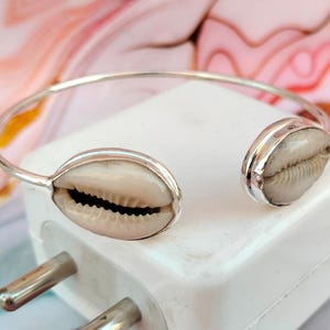 以下が含まれることがあります： 2つのカウリ貝のチャームが付いたシルバーのバングルブレスレット。ブレスレットはオープンタイプで、シンプルなデザインです。