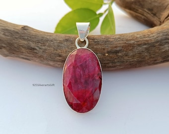 Könnte beinhalten: Ein roter Rubin-Edelstein-Anhänger in Sterling Silber. Der ovale Edelstein ist tiefrot und hat einen facettierten Schliff. Der Anhänger liegt auf einer Holzoberfläche.