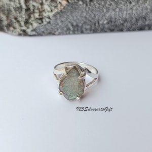 以下が含まれることがあります： 洋梨型のイリデッセント宝石をあしらったシルバーリング。リングはスプリットシャンクのデザインで、白い表面にセットされています。「925SilverartsGift」の文字がリングの下に見えます。
