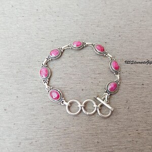 May include: A silver bracelet featuring oval-shaped, vibrant pink gemstones. The bracelet has a toggle clasp and is set against a neutral background. The text "925 SilverartsGift" is visible.