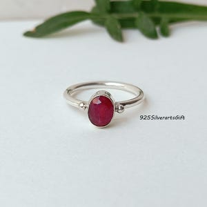 May include: A silver ring featuring a vivid red oval gemstone. The ring has a plain band with small decorative details either side of the gemstone. The text "925SilverartsGift" is visible on the right side of the image.