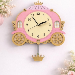 May include: A wall clock shaped like a pink carriage with gold wheels and a crown topper. The clock face is cream-coloured with black numbers and hands. A black pendulum hangs from the bottom, featuring a small gold crown with colourful gems.