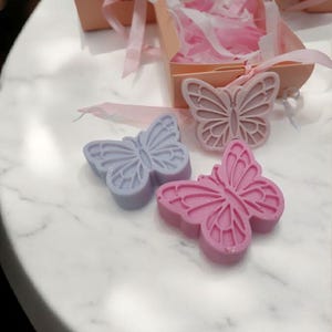 May include: Two butterfly-shaped soaps, one lavender and one pink, on a white marble surface. The soaps are in the shape of butterflies with detailed wings.