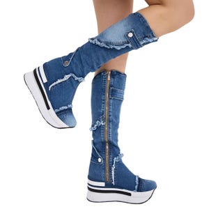 Könnte beinhalten: Kniehohe Stiefel aus blauem Denim mit ausgefransten Kanten und einem Umschlag mit silberfarbenem Druckknopf. Die Stiefel verfügen über einen durchgehenden goldenen Reißverschluss an der Seite und eine dicke weiße Plateausohle mit schwarzem Streifen.