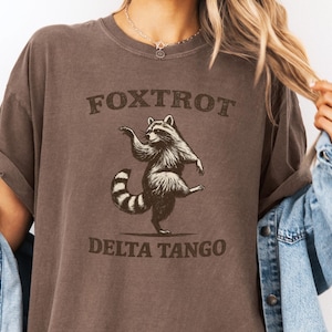 May include: Espresso-colored t-shirt with the text "FOXTROT DELTA TANGO" above and below a dancing raccoon illustration. The shirt is a Comfort Colors brand, style 1717.