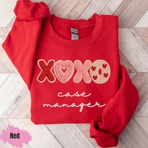Könnte beinhalten: Ein rotes Sweatshirt mit den gestickten Buchstaben "XOXO" und einem Herzdesign. Der Text "case manager" steht darunter in Schreibschrift. Das Sweatshirt liegt gefaltet auf einer Holzoberfläche.
