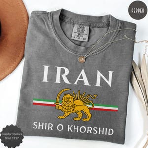 Pode incluir: Camiseta cinza Comfort Colors com a palavra "IRAN" em branco. Abaixo, um desenho de leão dourado com uma espada, sobre uma faixa vermelha, branca e verde. O texto "SHIR O KHORSID" está abaixo. Um colar de prata está na camiseta.