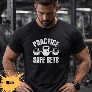 Könnte beinhalten: Schwarzes T-Shirt mit dem weißen Text "PRACTICE SAFE SETS" und einer Hantel- und Kettlebell-Grafik. Das Wort "Black" befindet sich in einem gelben Feld unten links. Das T-Shirt hat einen Rundhalsausschnitt.