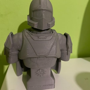 Pode incluir: Busto cinzento impresso em 3D de uma figura futurista blindada, com capacete e armadura nos ombros. O busto é montado numa base retangular com o texto "SUPER EARTH COMMANDER". A figura está sobre um fundo verde.