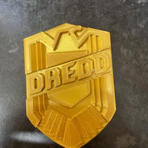 May include: A gold-colored, hexagonal badge with the word "DREDD" in bold, raised letters. The badge has a detailed, textured design with geometric shapes and lines. The overall aesthetic suggests a futuristic or sci-fi theme.