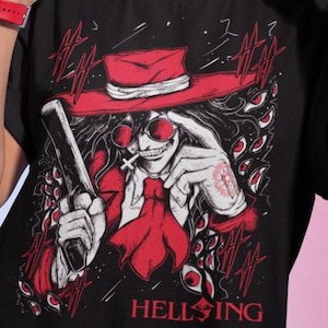 May include: Black t-shirt featuring a red and white graphic of a character with a hat, sunglasses, and a cigarette, holding a gun. The word "HELLSING" is printed in red. The person in the image is wearing gold hoop earrings and a red watch.