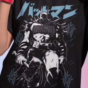 Puede incluir: Camiseta negra con un diseño gráfico de Batman y texto japonés en azul. El diseño es en blanco y negro. La persona que lleva la camiseta tiene pendientes de aro y pintalabios rojo.