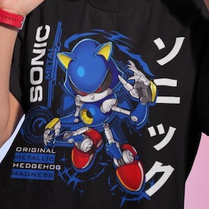 Peut inclure: T-shirt noir avec un motif de Sonic le Hérisson robotique bleu et argenté. Le t-shirt porte les inscriptions "SONIC METAL" et "ORIGINAL METALLIC HEDGEHOG MADNESS" en blanc et bleu. Du texte japonais est également présent.