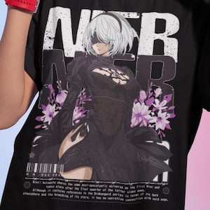 Könnte beinhalten: Schwarzes T-Shirt mit einer Grafik einer Figur aus dem Videospiel Nier: Automata, mit dem Titel des Spiels in weißen Blockbuchstaben. Das Design enthält lila Blumenakzente und Text am unteren Rand. Die Person trägt goldene Creolen.