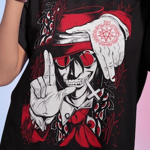 May include: Black t-shirt featuring a stylized illustration of a character with a top hat, sunglasses, and a cross. The design includes red accents and a hand with a pentagram tattoo. The person in the image is wearing gold hoop earrings and a red watch.