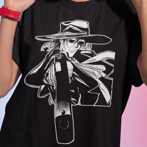 Puede incluir: Camiseta negra con un gráfico en blanco y negro de un personaje con sombrero y gafas de sol, sosteniendo un arma. La persona de la imagen lleva pendientes de aro dorados y un reloj rojo.