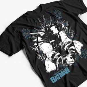 Op de afbeelding: Zwart t-shirt met een grafische illustratie van een stripfiguur. Het ontwerp is in wit en zwart, met blauwe accenten en de tekst "ABSOLUTE BATMAN". Het shirt heeft een ronde hals en korte mouwen.