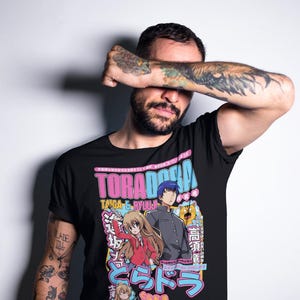 May include: Black t-shirt featuring anime characters and the text "TORADORA" in pink and blue. The design includes Japanese text and illustrations of two anime characters. The shirt is worn by a person with tattoos.