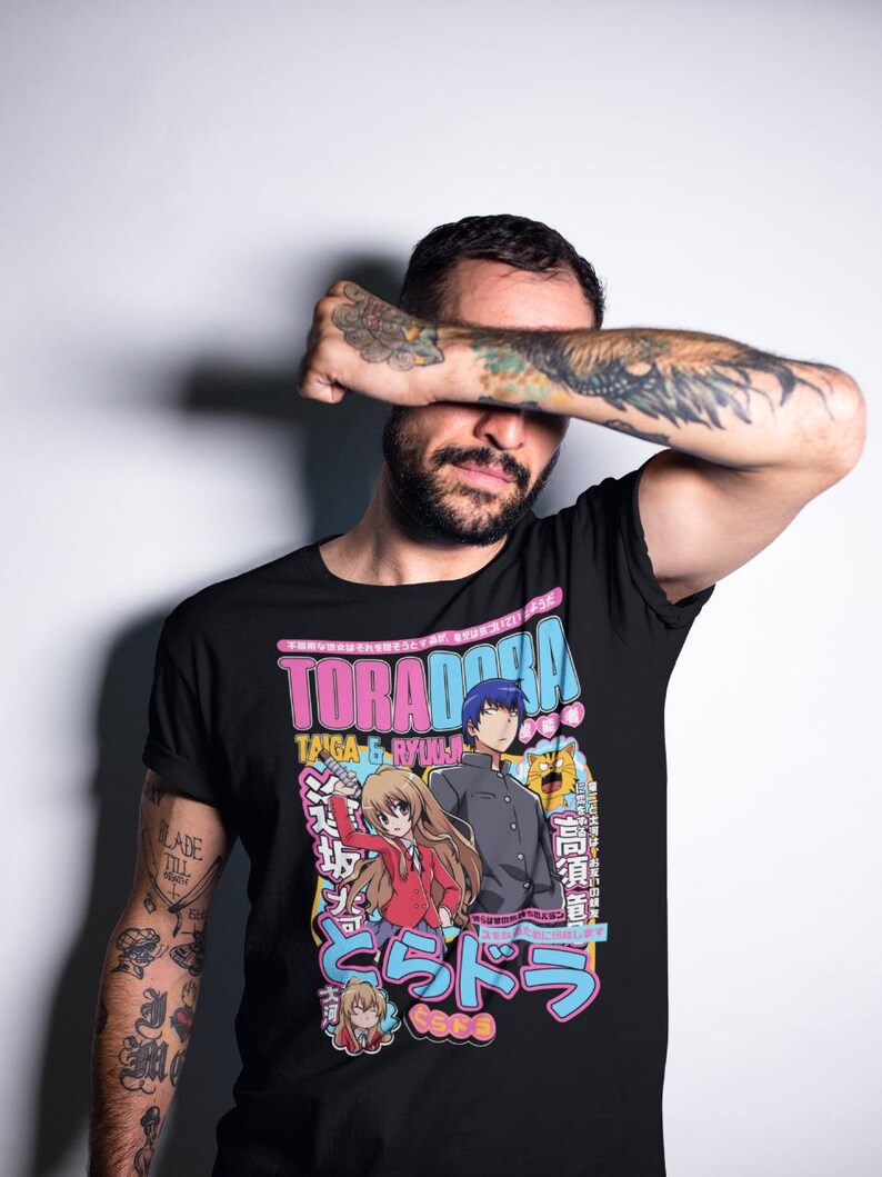 May include: Black t-shirt featuring anime characters and the text "TORADORA" in pink and blue. The design includes Japanese text and illustrations of two anime characters. The shirt is worn by a person with tattoos.