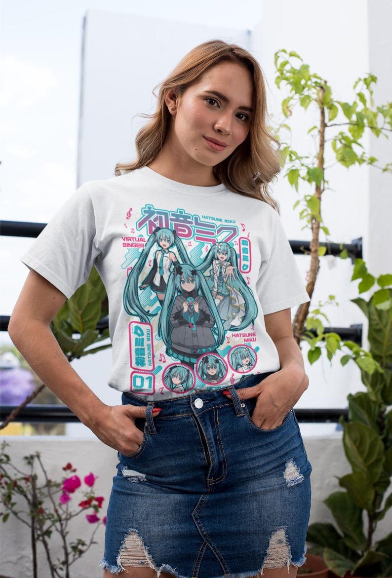 Puede incluir: Camiseta blanca con un gr&aacute;fico colorido de una cantante virtual con largo cabello turquesa. El dise&ntilde;o incluye texto japon&eacute;s y las palabras en ingl&eacute;s "Virtual Singer". Llevada por una persona en mezclilla azul.