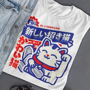 May include: White t-shirt with a Japanese lucky cat graphic in blue and red. The shirt features Japanese text and is paired with a denim jacket, sunglasses, and a beaded bracelet. A casual, stylish look.