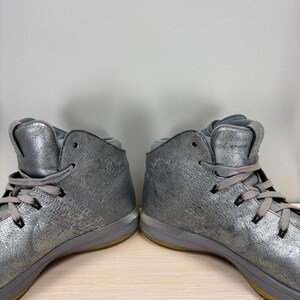 May include: Pair of silver high-top sneakers with gray laces and a textured metallic finish. The shoes have a gray sole with a yellow accent and are displayed on a light wood surface.