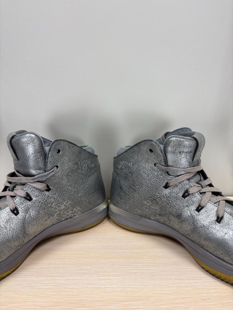 May include: Pair of silver high-top sneakers with gray laces and a textured metallic finish. The shoes have a gray sole with a yellow accent and are displayed on a light wood surface.
