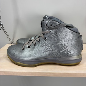 May include: Metallic silver high-top athletic shoe with gray laces and a gray sole. The shoe features a silver Nike swoosh and a Jumpman logo. The sole is approximately 1 inch thick.