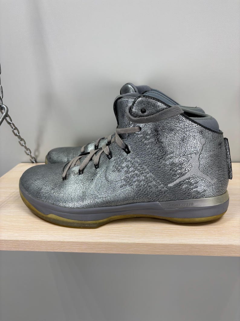 May include: Metallic silver high-top athletic shoe with gray laces and a gray sole. The shoe features a silver Nike swoosh and a Jumpman logo. The sole is approximately 1 inch thick.