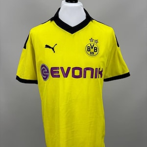 May include: A yellow soccer jersey with black trim and a black collar. The jersey features the Puma logo, the text "EVONIK" in purple, and a team emblem. The jersey is displayed on a mannequin.