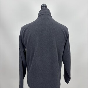 May include: A dark grey fleece jacket with a high collar and a zip closure. The long-sleeved jacket is displayed on a mannequin against a plain background. The jacket has a simple, classic design.