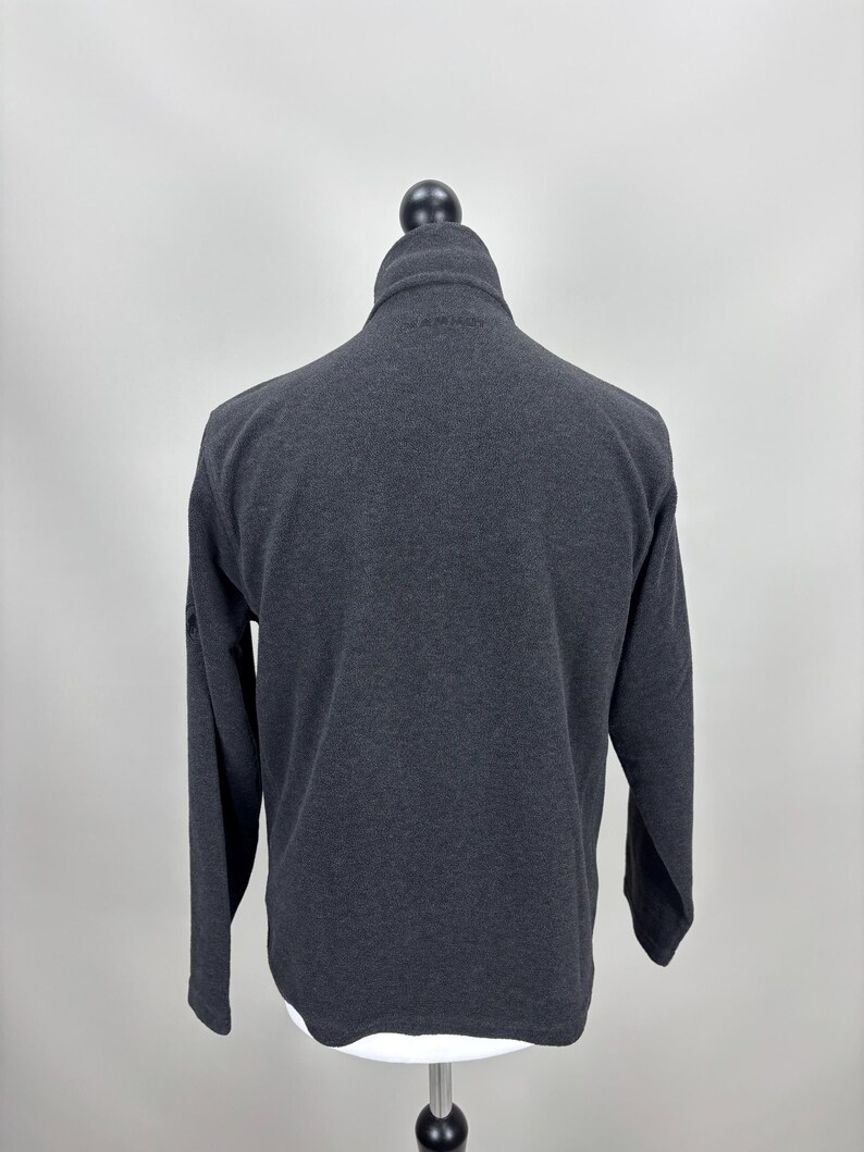 May include: A dark grey fleece jacket with a high collar and a zip closure. The long-sleeved jacket is displayed on a mannequin against a plain background. The jacket has a simple, classic design.