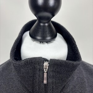 May include: A white ceramic mug with a black handle and the text "But First, Coffee" in a bold, black font. The mug is set against a blurred background. The mug appears to be approximately 10 cm tall.