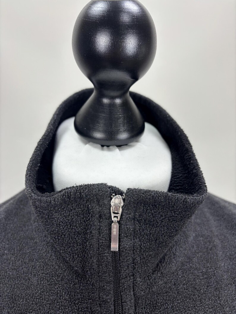 May include: A white ceramic mug with a black handle and the text "But First, Coffee" in a bold, black font. The mug is set against a blurred background. The mug appears to be approximately 10 cm tall.