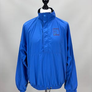 May include: Bright blue windbreaker jacket with a high collar and a quarter-zip closure. The jacket features a small logo on the chest and a drawstring at the hem. The sleeves are long and have elastic cuffs. The jacket is displayed on a mannequin.